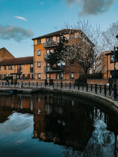 A view of a residential waterfront area in Enfield Lock featuring a three-story brick apartment building with balconies, surrounded by leafless trees and a paved promenade with black metal railings. T