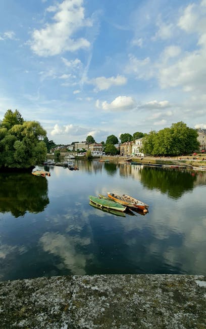 A tranquil river scene featuring calm, reflective water with gentle ripples, bordered by lush green trees and well-maintained residential buildings lining the far bank. Several small boats in green an
